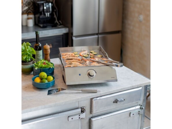 Mise en situation d'une grillade de crevettes à la plancha sur le plan de travail d'une cusine intérieure à côté d'herbes aromatiques, de citrons, d'huile olive et de poivre