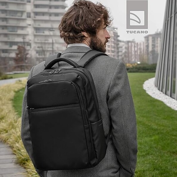 Homme brun et barbu portant un costume gris les mains dans les poches portant le sac à dos Tucano sur ses épaules en extérieur devant des immeubles un jour de ciel gris