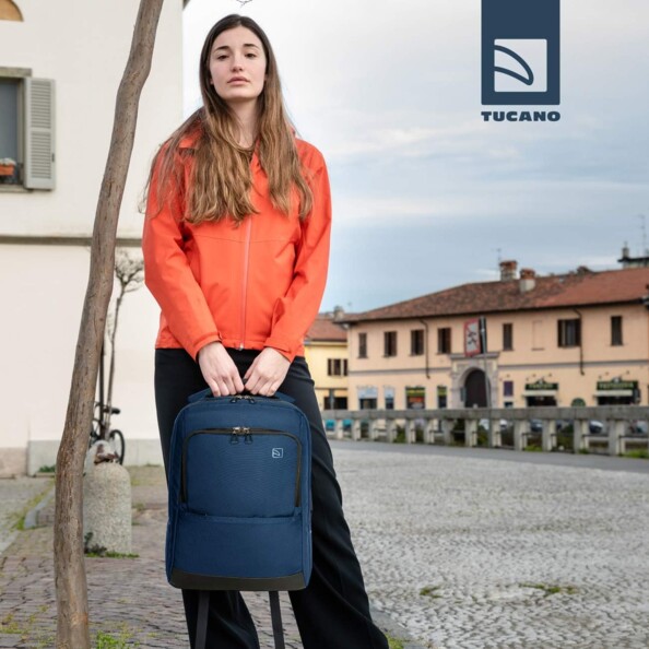 Jeune femme en veste orange et bas noir debout à côté d'un arbre, le sac Tucano dans les mains dans une rue pavé devant des bâtiments typiques de village