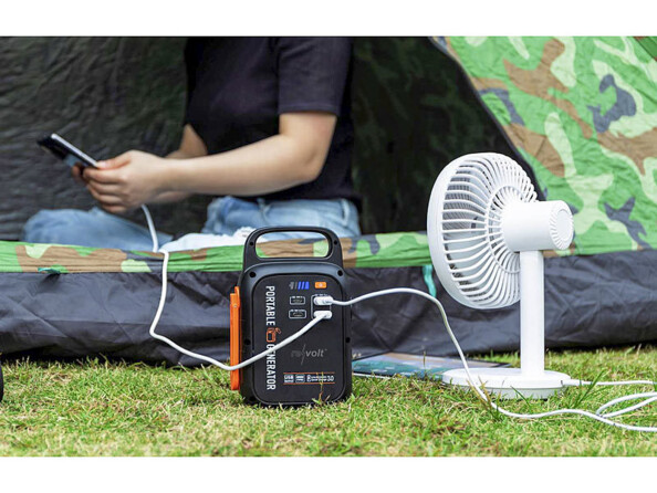 Femme en tailleur assise dans une tente au motif camouflage avec un téléphone en main branché à la batterie de secours PB-100.k, elle-même branchée à un mini ventilateur USB blanc