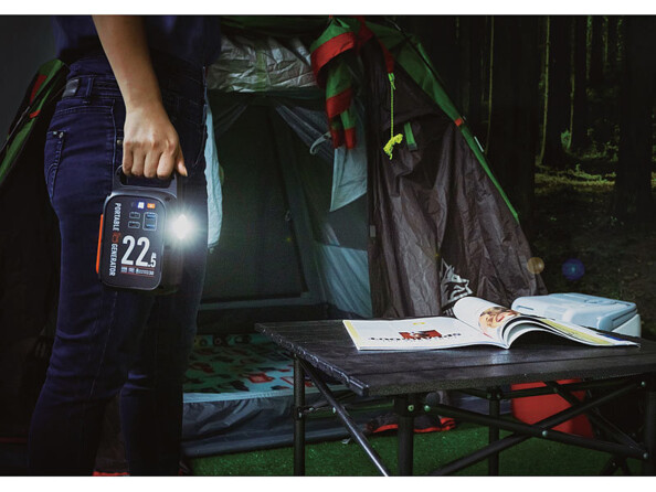 Personne debout devant une tente ouverte et une table sur laquelle se trouve des documents dans le noir portant dans sa main droite la batterie d'appoint lithium-ion dont la lampe de poche LED est allumée