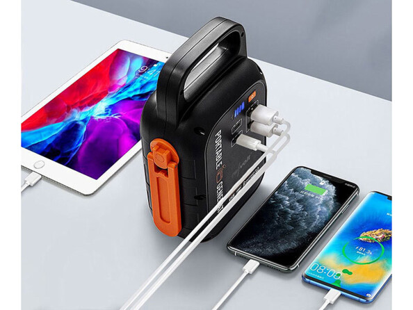 Vue en plongée sur la batterie d'appoint à dynamo coloris orange et noir posée sur un meuble et branchée à deux smartphones et une tablette allumés par le biais de trois câble de chargement USB et des ports USB-A et USB-C de l'appareil