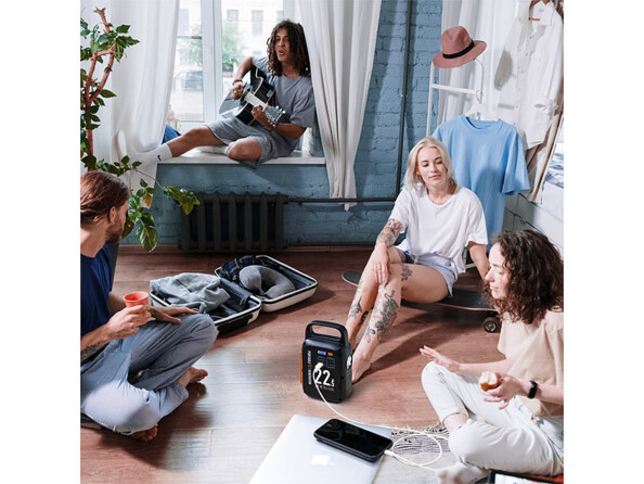 Quatre amis dont 2 femmes et 2 hommes dans une chambre au style bohème assis par terre ou sur le rebord de fenêtre avec au milieu d'eux la batterie externe PB-100.k entrain de recharger un téléphone portable sur une commode blanche