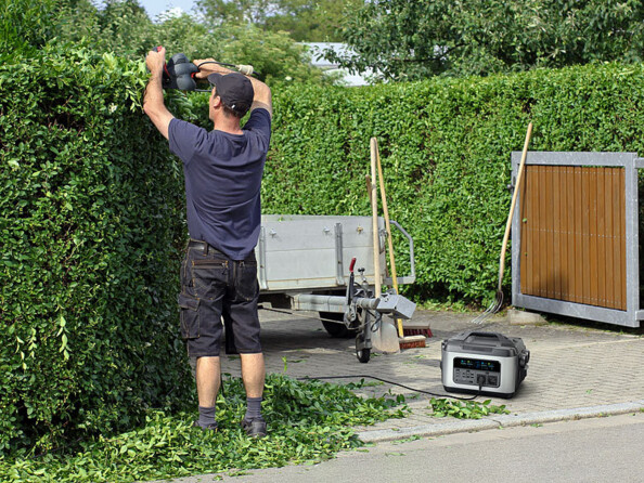 Homme entrain de tailler une haie de conifères avec un taille-haie alimenté par la batterie d'appoint LiFePO4 100 Ah avec remorque et outils de jardinage à côté