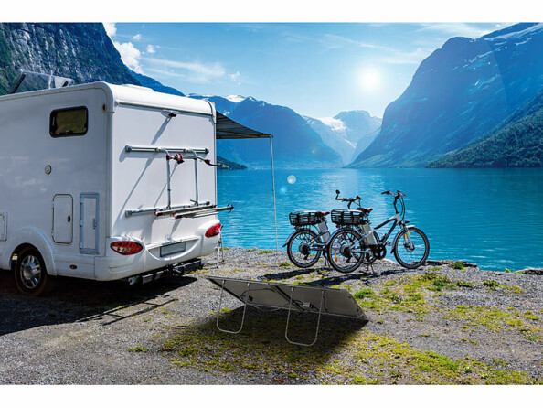 Mise en situation du panneau solaire déplié vers le soleil au bord d'un lac de montagne derrière un camping-car garé et de vélos posés devant le plan d'eau avec montagnes en relief en arrière-plan