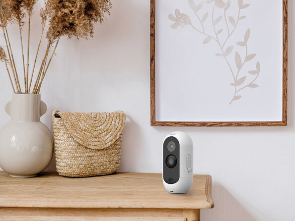 Caméra de sécurité ovale blanche et noire posée à la verticale sur un meuble en bois beige à côté d'un tableau, d'une besace et d'un pot de fleur décoratif dans les mêmes tons