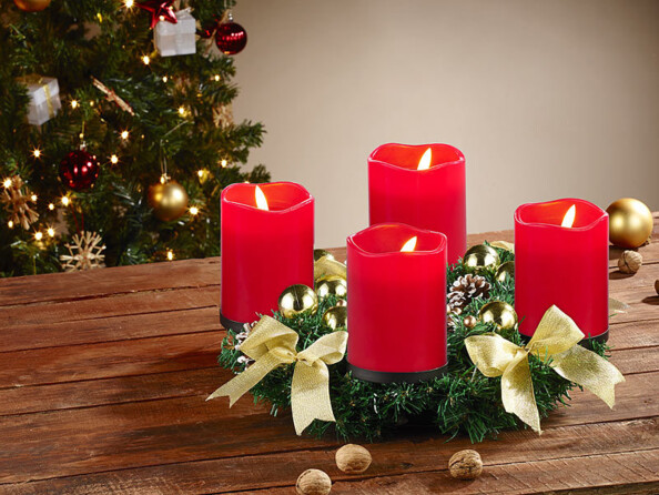 Couronne de l'Avent coloris doré avec 4 bougies LED coloris rouge posée sur une table de salon en bois à côté de noix entières devant un sapin de Noël décoré de guirlandes lumineuses et décorations de Noël