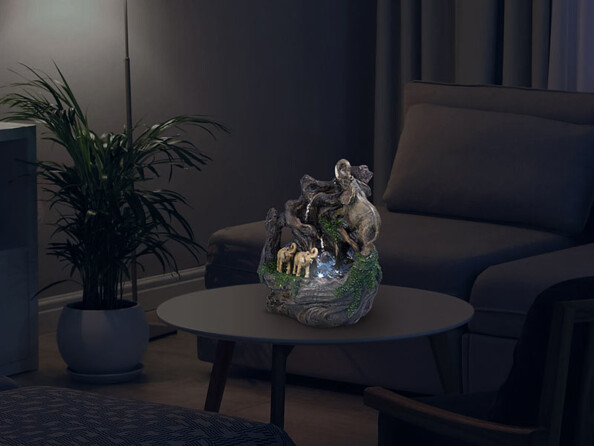 Mise en situation de la fontaine à eau avec éléphants décoratifs dans une cascade créée dans un arbre posée sur une table basse dans un salon avec canapé, fauteuil, tapis et plante verte, le tout dans l'obscurité