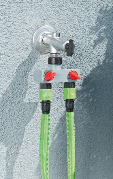 Utilisation du répartiteur d'eau branché à une arrivée d'eau extérieure sortant de la façade d'une maison et fournissant de l'eau à deux tuyaux d'arrosage verts en même temps