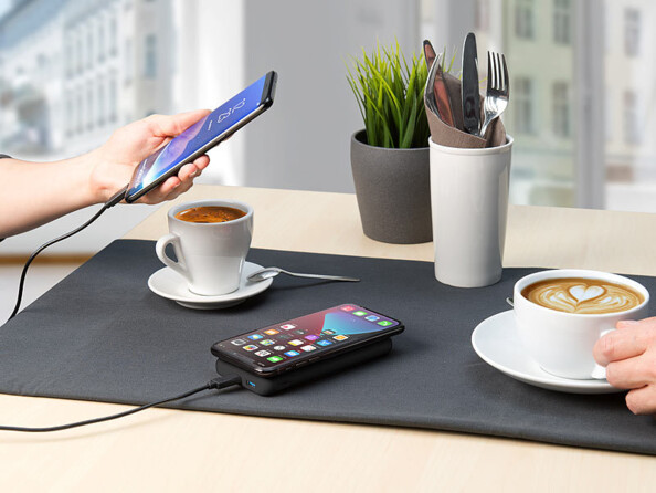 Main d'une personne tenant un téléphone portable allumé branché à une batterie externe posée sur un set de table à côté de couverts et de deux tasses de café rechargeant également un second téléphone mobile par induction