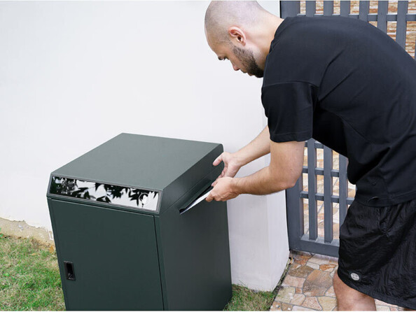 Homme chauve en short et t-shirt noir glissant une enveloppe dans la boîte aux lettres devant une propriété