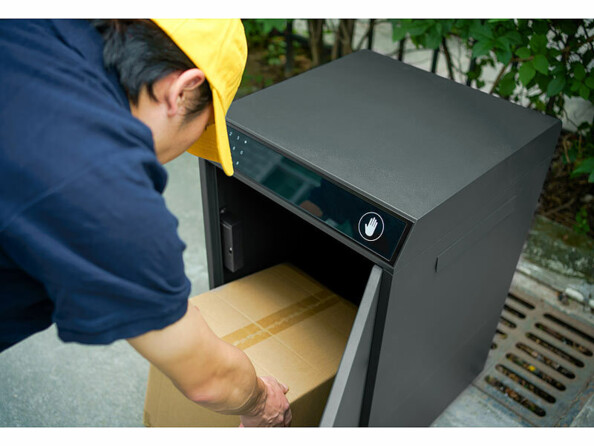 Livraison de courrier par un facteur en tenue noire avec casquette jaune qui glisse le colis dans la boîte aux lettres posée au sol dans un jardin