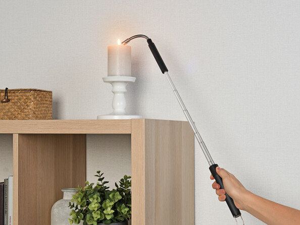 Personne tenant dans sa main le briquet télescopique Pearl étendu en train d'allumer la flamme d'une bougie installée dans son bougeoir sur un meuble en bois à cases