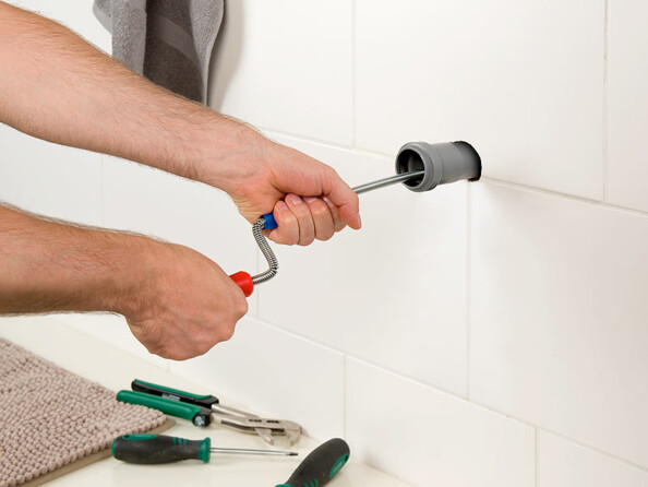 Plombier entrain de déboucher un tuyau d'évacuation d'eau sale sortant d'un mur carrelé à deux mains à l'aide d'une spirale de nettoyage à manivelle avec tournevis, pince et autres outils à côté