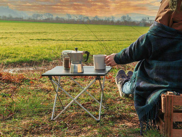 Personne en veste et capuche assise sur une cagette en palette devant un champ sur lequel se couche le soleil, tenant une tasse posée sur la table de camping Semptec dans sa main gauche à côté d'une cafetière Cucina Di Modena