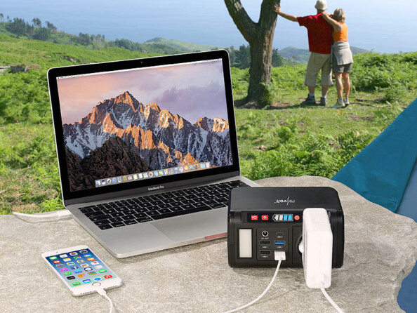 Mise en situation de la batterie nomade HSG-240 posée sur une table de camping en extérieur entrain de recharger un ordinateur portable via sa prise 230 V et un smartphone via son port USB avec en arrière-plan un couple qui se tient à un arbre