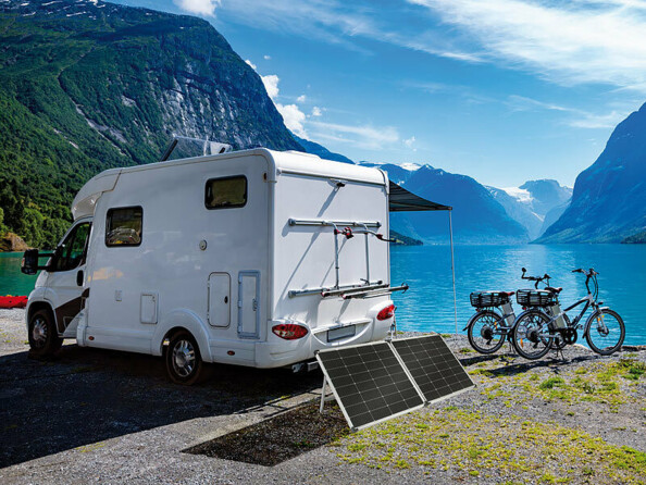 Panneau solaire déplié au sol derrière un camping-car blanc et à côté de deux vélos, devant un lac avec en arrière-plan les montagnes, le tout pendant une journée ensoleillée