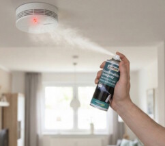 ne personne vaporise le spray de test STANGER vers un détecteur de fumée fixé au plafond d'un salon, déclenchant le voyant lumineux rouge de l'appareil."