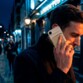 Homme trentenaire cheveux bruns avec manteau téléphonant en extérieur de nuit dans la rue avec un smartphone doré sur lequel est fixé une batterie externe de la même couleur par induction