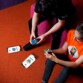 Concours de résolution rapide de Rubiks cube entre une jeune femme en t-shirt orange assise par terre et un jeune homme habillé en noir sur un pouf rose sur un sol en moquette orange
