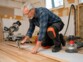 Homme agenouillé installant un parquet avec genouillères, marteau et ponceuse orbitale