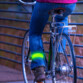 Personne en jean de dos faisant du vélo devant un bâtiment en briques rouges avec la bande lumineuse et réfléchissante couleur fluo à la cheville
