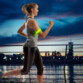 Femme châtain blonde les cheveux attachés et en tenue de sport courant sur un pont le soir dans l'obscurité avec le bracelet lumineux LED SlapLit NiteIze porté autour du biceps