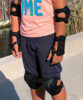 Enfant en t-shirt rose et short bleu équipé de coudières, protège-poignets et genouillères noires près de l’eau