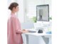Mise en situation d'une jeune femme brune en blazer rose pâle entrain de travailler à son bureau debout sur ordinateur dans un setup informatique moderne