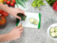 Mise en situation d'une personne découpant une courgette sur une planche à découper blanche et verte sur un plan de travail en granit à côté de légumes