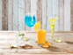 Mise en situation de deux pailles droites et deux pailles courbées dans 4 verres différents dans lesquels sont servis plusieurs cocktails sur une table de jardin en bois brut clair