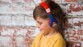 Petit garçon châtain souriant adossé contre un mur en briques rouges portant un t-shirt jaune moutarde et le casque audio Super Mario OTL Technologies sur les oreilles