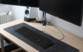 Mise en situation du clavier filaire et hub USB 2 ports 2 en 1 sur un tapis noir en velours sur un bureau en bois, branché à un PC éteint à côté d'une enceinte et d'un radiateur