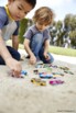 2 petits garçons en t-shirt et jean assis par terre dans un parc en extérieur entrain de jouer aux petites voitures dans le sable avec des bolides de toutes les couleurs