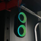 Enceinte bluetooth sans fil d'extérieur TOOGO M Ryght suspendue à une barre métallique d'un appareil de musculation par le biais de ses lacets bleus avec LED allumées en vert dans l'obscurité de la salle