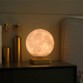 Lampe décorative MoonFlight avec lune 3D en vol stationnaire au-dessus d'une base posée sur une tablette de nuit en bois chic sur laquelle sont posés des objets dorés à côté d'un lit