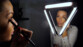 Femme souriante se maquillant les pommettes avec un gros pinceau devant le miroir avec la lampe pour miroir Wellys disposée en forme de pont sur le miroir par bande adhésive