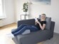 Mise en situation de l'oreiller en forme de trapèze utilisé comme repose-pieds par une femme allongée sur le canapé entrain de lire dans le salon