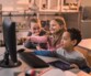 Trois jeunes enfants souriants dans une salle de classe regroupés devant un écran d'ordinateur avec clavier et souris filaire posés sur un bureau devant une tablette et des fournitures d'école montrant du doigt le contenu à l'écran