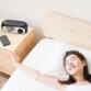 Vue en plongée d'une femme asiatique allongée dans un lit blanc et bois clair souriant et s'étirant les bras au réveil avec à côté du lit une commode du même coloris sur laquelle est posé un smartphone et le réveil 2 en 1 à surface miroir affichant 8:00