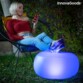 Adolescente blonde aux cheveux courts en jean et crop top à une manche rouge regardant son téléphone le sourire aux lèvres assise dans un fauteuil de jardin dans le noir avec les pieds étendus et posés sur le pouf gonflable LED illuminé d'un bleu indigo