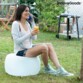 Jeune femme brune en gilet, débardeur, short et basket assise sur le pouf gonflable éteint avec un verre de jus de fruit à la main et un téléphone portable dans l'autre dans un jardin