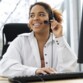 Jeune femme métisse souriante au téléphone avec un client assise devant un PC avec la main droite sur le clavier filaire