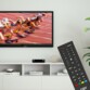 Mise en situation du décodeur TNT DVB-T2 sur un meuble blanc moderne sous une télévision affichant une course d'athlétisme avec au premier plan la télécommande fournie en accessoire
