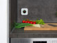 Détecteur de gaz carré blanc avec écran et voyant LED fixé à la crédence d'un plan de travail de cuisine devant une planche sur laquelle se trouve divers légumes et un couteau, au-dessus d'un lave-vaisselle encastré