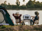 Deux compagnons détendus posés le long d'un plan d'eau sur leur chaise de camping avec la batterie nomade et convertisseur solaire HSG-1200 posée sur leur table et tente au premier plan