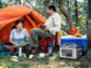 Couple en camping assis devant une tente orange montée, alimentation leur glacière bleu et blanche par le biais de la batterie nomade HSG-1200