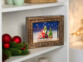 Cadre décoratif de Noël "Père Noël et cadeaux" posé sur une étagère blanche à côté d'une décoration de Noël avec branches de sapin et boules rouge brillant