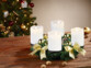Couronne de l'Avent coloris doré avec 4 bougies LED blanches posée sur une table de salon en bois à côté de noix entières devant un sapin de Noël décoré de guirlandes lumineuses et décorations de Noël