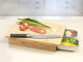 Mise en situation de la planche à découper en bambou avec tomate et oignons découpés posés à côté d'un couteau de chef sur la planche et ingrédients découper dans un des deux bacs en acier avec les restes et épluchures dans le second